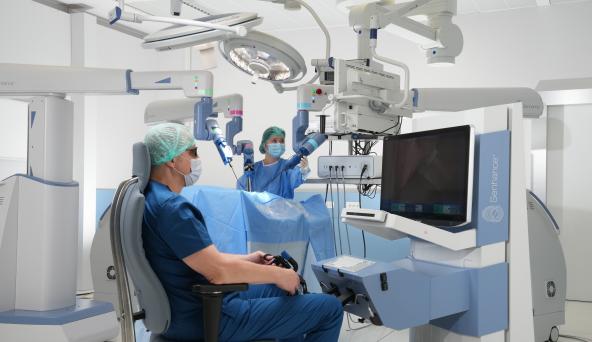 A surgeon sitting in a chair while another surgeon is in the background with a robot